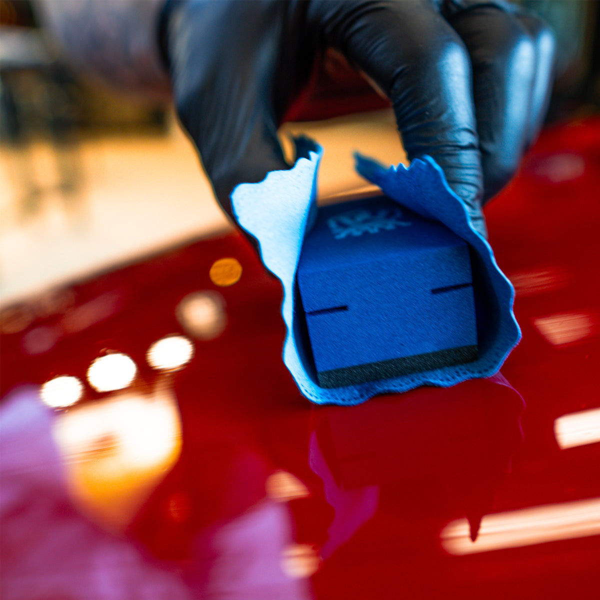 Blue foam block with a black square in the center, held by a gloved hand against a blurred red car background.
