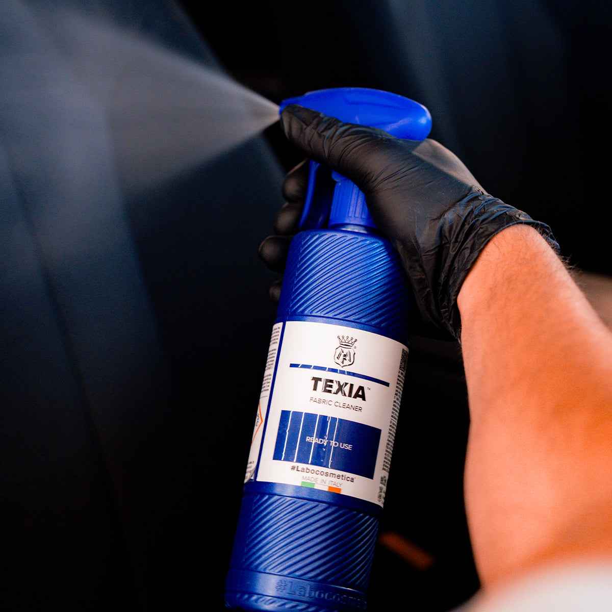 Hand holding a blue spray bottle labeled 'TEXIA' against a dark background