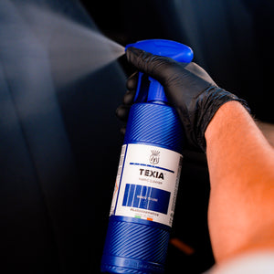 Hand holding a blue spray bottle labeled 'TEXIA' against a dark background