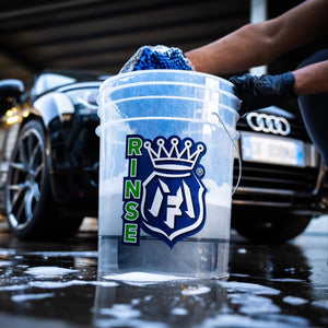Clear bucket with 'RINSE' logo held by a person at a car wash with Audi cars in the background.