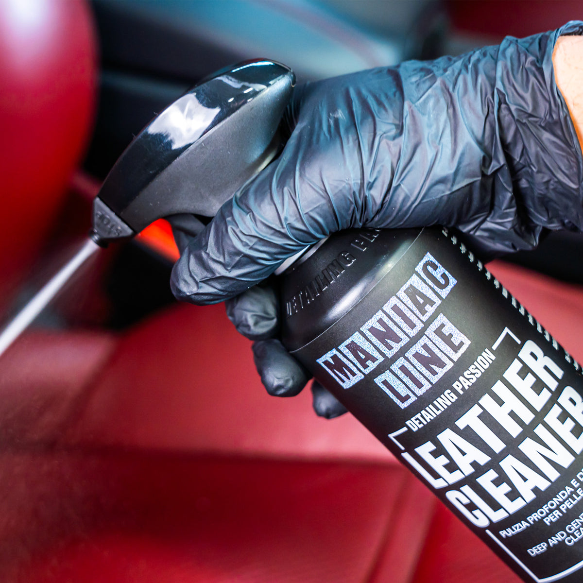 Hand wearing a black glove holding a can of leather cleaner with a red car interior in the background.