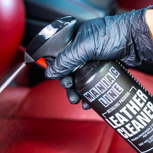 Hand wearing a black glove holding a can of leather cleaner with a red car interior in the background.