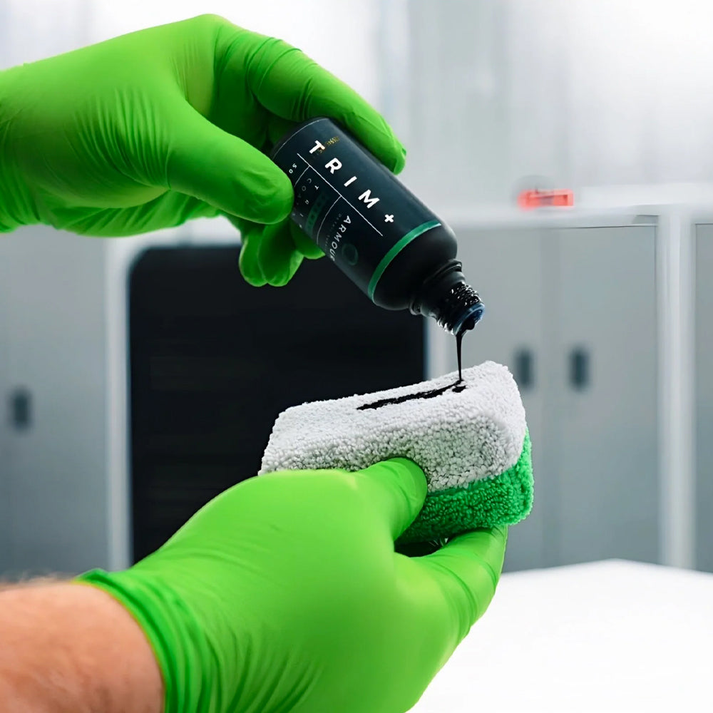 Person in green gloves applying a black liquid from a 'TRIM' bottle onto a white cloth.