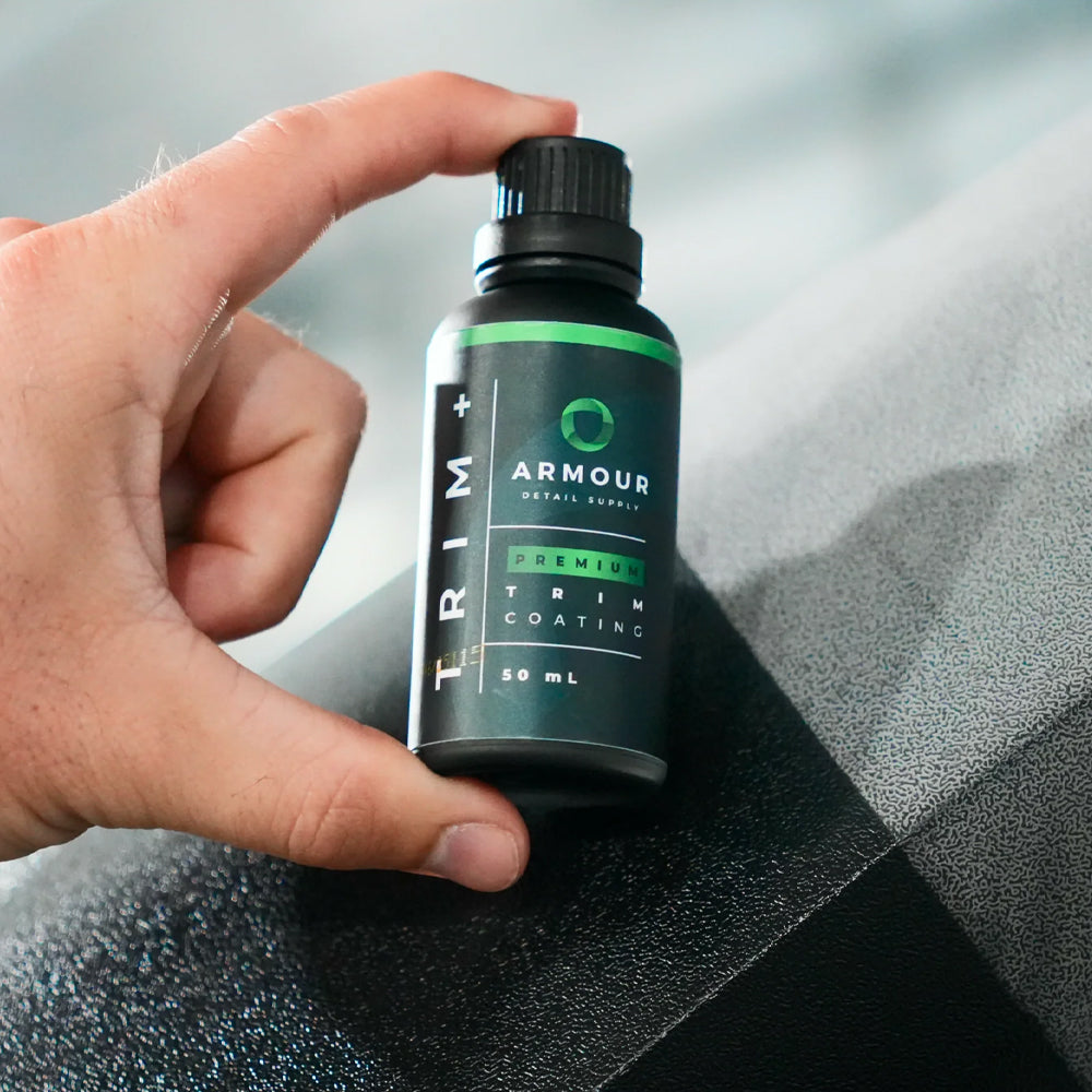 Hand holding a bottle of Armour Trim+ Premium Trim Coating against a car interior background