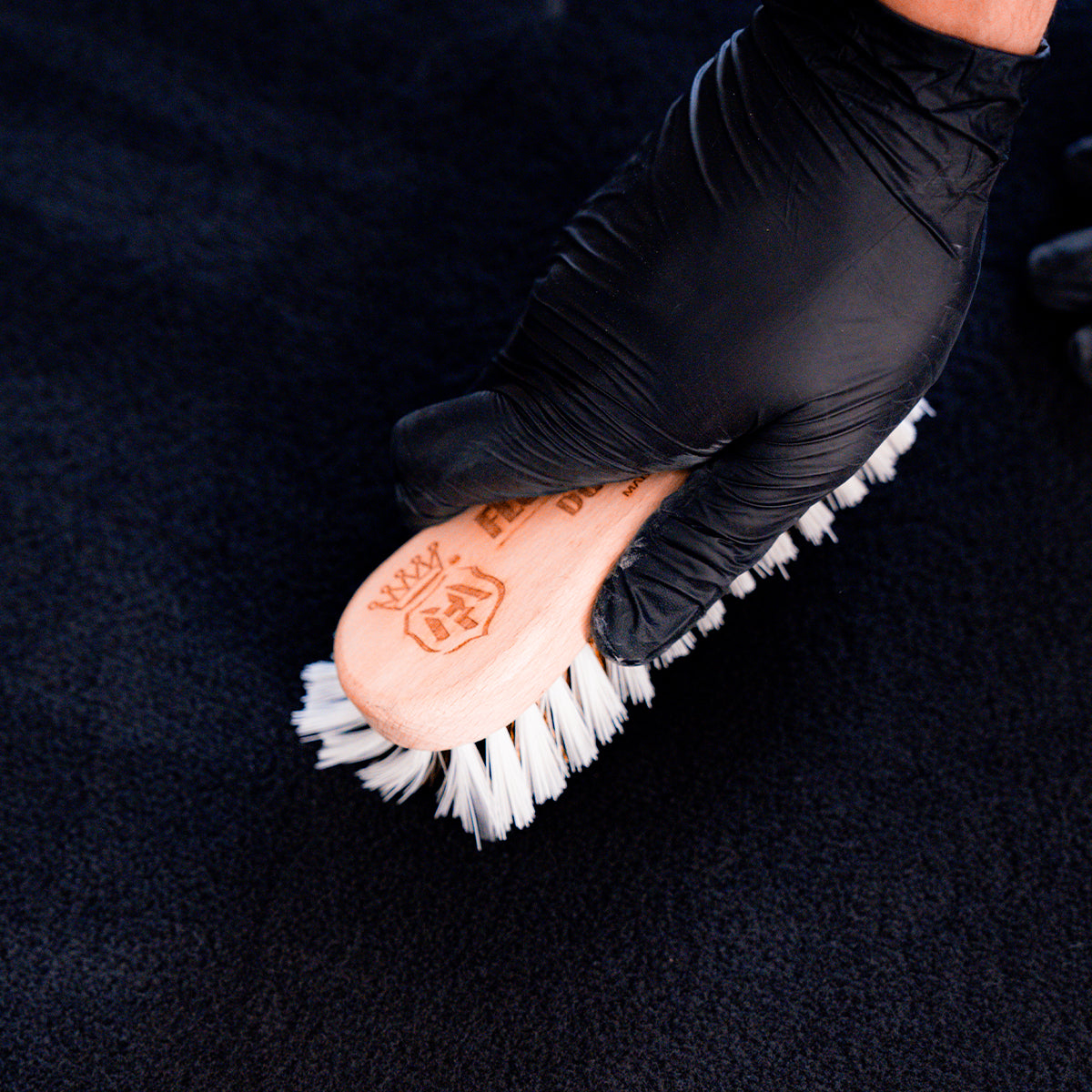 Person wearing black gloves holding a pink brush with white bristles on a dark background