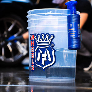 Clear bucket with 'WHEELS' logo held by a person at a car wash with Audi cars in the background.